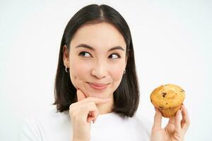 vicino su ritratto di asiatico donna con pensieroso sorridente viso, detiene cupcake, pensa per mangiare Pasticcino, bianca sfondo foto