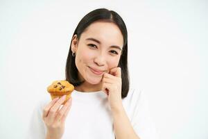 vicino su ritratto di asiatico ragazza con cupcake, pensiero e sorridente, mangiare dolce, frenata dieta, bianca sfondo foto