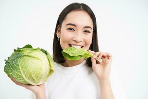 vicino su ritratto di asiatico donna, pungente lattuga, mangiare verde cavolo e sorridente, bianca sfondo foto