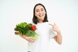 Immagine di contento coreano donna, puntamento a la verdura, sorridente, consigliare mangiare salutare crudo cibo, verde dieta concetto, bianca sfondo foto