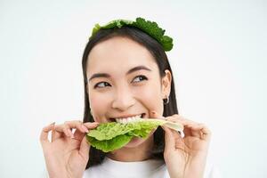 vicino su ritratto di sorridente asiatico donna mangiare lattuga, gli amori cavolo, vegetariano gode crudo biologico cibo, bianca sfondo foto