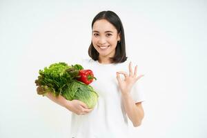 ritratto di sorridente asiatico donna, Tenere mazzo di verdure e fa va bene cartello, raccomanda verde nutriente cibo consegna app, bianca sfondo foto