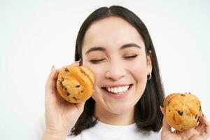 vicino su ritratto di asiatico donna, Spettacoli Due cupcakes vicino viso e sorrisi, piace Pasticcino, gode mangiare forno, bianca sfondo foto