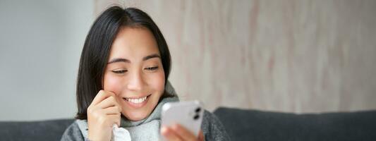 ritratto di coreano donna si sente malato, Tenere smartphone, chiamata medico gp per ottenere prescrizione, catturato freddo, soggiorno a casa, utilizzando mobile Telefono foto