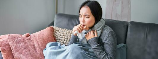 asiatico donna tosse, sensazione malato, seduta sotto coperta con scard nel freddo appartamento, Salvataggio su riscaldamento Questo inverno, attraente influenza foto