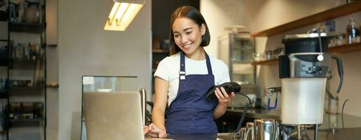 sorridente asiatico ragazza barista, Lavorando nel bar, servendo cliente, guardare a il computer portatile mentre utilizzando carta lettore per ricevere i soldi per ordine, vendita caffè foto