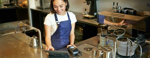 sorridente asiatico ragazza barista Lavorando nel bar a contatore, in lavorazione ordini, utilizzando pos terminale ricevente pagamento per caffè foto