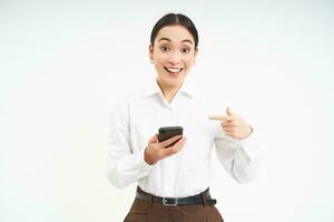 sorridente coreano aziendale donna, puntamento a smartphone e guardare felice, mostrando su mobile Telefono app, in piedi al di sopra di bianca sfondo foto