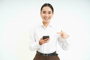 sorridente coreano aziendale donna, puntamento a smartphone e guardare felice, mostrando su mobile Telefono app, in piedi al di sopra di bianca sfondo foto