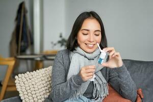 donna guardare a sua spray a partire dal dolorante gola, trattamento sua freddo, soggiorno a casa malato, sensazione meglio dopo influenza, seduta nel sciarpa a casa nel vivente camera foto