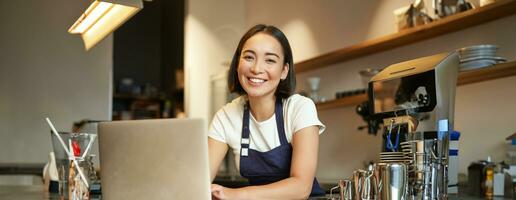 entusiasta asiatico ragazza nel bar uniforme, barista lavoratore con computer portatile, guardare contento e sorpreso a telecamera foto