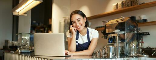 ritratto di sorridente coreano donna, barista nel caffè negozio, in piedi a contatore con computer portatile, sorridente e guardare fiducioso, lavoratore autonomo femmina imprenditore nel sua proprio caffè negozio foto