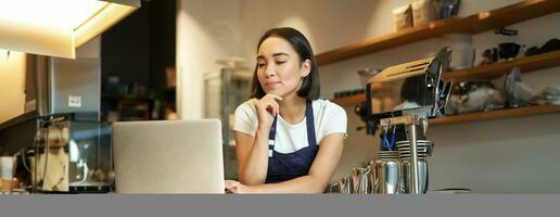 ritratto di sorridente coreano donna, barista nel caffè negozio, in piedi a contatore con computer portatile, sorridente e guardare fiducioso, lavoratore autonomo femmina imprenditore nel sua proprio caffè negozio foto