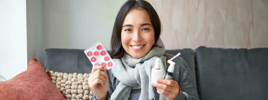 ritratto di contento sorridente asiatico ragazza mostrando farmaco, dolorante gola spray e farmaci a partire dal influenza o freddo, soggiorno a casa, indossare caldo Abiti foto