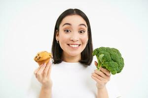 ritratto di coreano femmina modello, Tenere Cupcake e broccoli, mangiare salutare cibo, la scelta bene dieta per salutare corpo, bianca sfondo foto