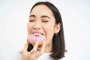 vicino su di sorridente lieta asiatico donna, mangia smaltato rosa ciambella, gode mangiare gustoso ciambella, bianca studio sfondo foto