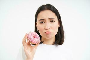 irritato singhiozzando ragazza cant mangiare Rifiuto cibo, Spettacoli delizioso smaltato ciambella, vuole per mordere, bianca sfondo foto