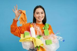 entusiasta asiatico donna sembra contento mentre pulisce casa a partire dal plastica sciupare, raccoglie sciocchezze, ordinamento spazzatura, blu sfondo foto