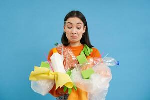 confuso asiatico donna detiene plastica sciupare, raccoglie spazzatura per riciclare, impara Come per ordinare diverso tipi di sciocchezze, concetto di eco sostenibile vita, blu sfondo foto