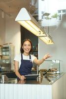 contento asiatico ragazza barista lavori nel caffè negozio, onde a cliente, in lavorazione ordini dietro a contatore nel bar, indossa blu grembiule foto