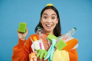 entusiasta asiatico ragazza mostrando pulizia spugna, Tenere vuoto plastica bottiglie, spazzatura per raccolta differenziata, ordinamento sua domestico spazzatura, blu sfondo foto