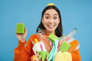 sorridente entusiasta femmina eco attivista, Tenere pulizia spugna, vuoto plastica bottiglie e spazzatura, ordinamento rifiuto per raccolta differenziata, blu sfondo foto