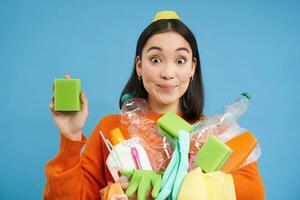 sorridente entusiasta femmina eco attivista, Tenere pulizia spugna, vuoto plastica bottiglie e spazzatura, ordinamento rifiuto per raccolta differenziata, blu sfondo foto