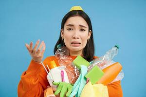 frustrato asiatico donna pianto, Tenere carichi di vuoto plastica bottiglie e spazzatura per raccolta differenziata, guardare stressato, blu sfondo foto