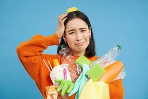 asiatico donna complicato di raccolta differenziata, Tenere mucchio di plastica sciupare, graffi testa, sembra infastidito, blu sfondo foto