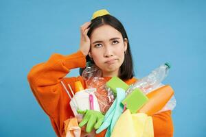 asiatico donna complicato di raccolta differenziata, Tenere mucchio di plastica sciupare, graffi testa, sembra infastidito, blu sfondo foto