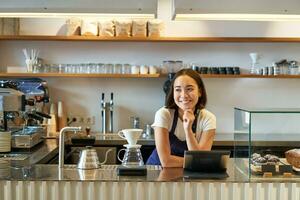 bar e piccolo attività commerciale. sorridente asiatico barista nel grembiule, in piedi dietro a contatore nel caffè negozio, Lavorando con pos terminale e filtro birra kit foto