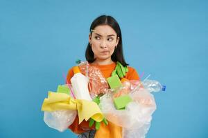 eco donna Tenere plastica bottiglie e raccolta differenziata sciupare, pensando, guardare confuso, ordinamento spazzatura in diverso bidoni, blu sfondo foto