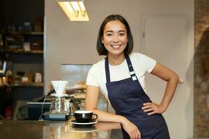 ritratto di asiatico sorridente barista, ragazza servendo caffè, in piedi vicino bar contatore nel grembiule, preparazione cappuccino per cliente foto