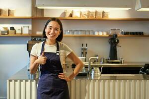 sorridente ragazza alunno Lavorando mezza giornata nel bar, barista Spettacoli pollici su, indossa grembiule, sta vicino caffè negozio contatore foto