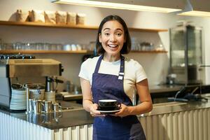ritratto di sorridente asiatico femmina barista, fabbricazione caffè, Tenere tazza di tè e assunzione esso per bar cliente, indossare grembiule, in piedi vicino contatore foto