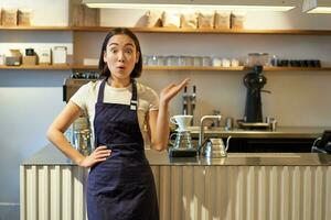 Immagine di eccitato asiatico femmina barista, guardare sorpreso, puntamento a smth nel bar, in piedi vicino caffè negozio contatore foto