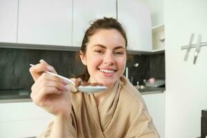 ritratto di sorridente bellissimo donna, alimentazione voi cucchiaio di cereali con latte, mangiare prima colazione e guardare felice, godendo sua mattina, indossare accogliente accappatoio foto