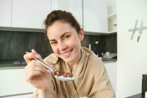 bellissimo sorridente donna, Tenere cucchiaio di cereali con latte, alimentazione voi e guardare con amore e cura, seduta nel cucina, avendo prima colazione nel mattina foto