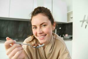 ritratto di bellissimo donna a casa, mangiare sua colazione, Tenere cucchiaio, pendente su piano di lavoro e avendo cereali con latte foto