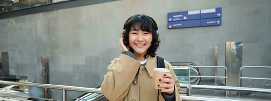 bellissimo sorridente coreano ragazza, bevande porta via caffè, ascolta musica nel cuffia, indossa trincea e zaino, sta su treno stazione, in viaggio foto