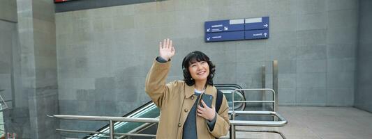 ritratto di carino sorridente asiatico ragazza nel cuffia, sta su strada con smartphone, onde mano a qualcuno, dice ciao, saluta amico foto