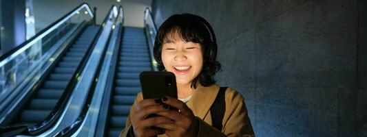 ridendo carino coreano ragazza su scala mobile, andando giù per tubo, metropolitana o la metropolitana, utilizzando mobile Telefono, Guardando smth divertente su smartphone App foto
