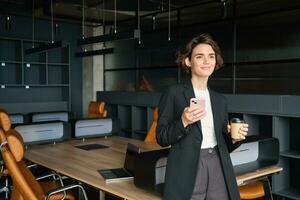 Immagine di donna nel conferenza camera. donna d'affari nel completo da uomo, bevande sua caffè e detiene smartphone, in attesa per incontro per inizio nel ufficio foto