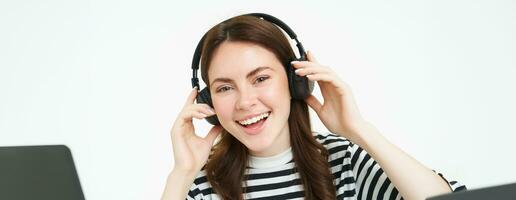 ritratto di donna, sorridente, indossare senza fili cuffia, ascoltando musica, studiando nel auricolari, in piedi isolato al di sopra di bianca sfondo foto