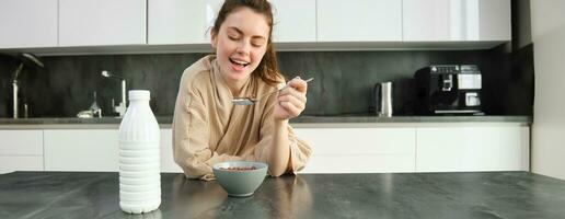 ritratto di bellissimo giovane e salutare donna nel accappatoio mangia sua prima colazione nel cucina, ha cereali con latte e sorridente foto