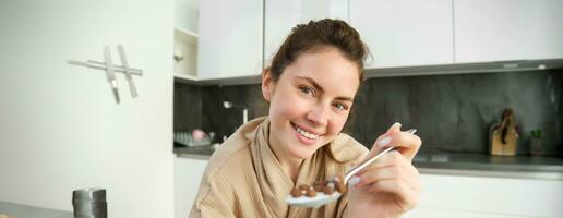 bellissimo sorridente donna, Tenere cucchiaio di cereali con latte, alimentazione voi e guardare con amore e cura, seduta nel cucina, avendo prima colazione nel mattina foto