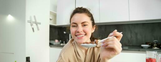 ritratto di sorridente bellissimo donna, alimentazione voi cucchiaio di cereali con latte, mangiare prima colazione e guardare felice, godendo sua mattina, indossare accogliente accappatoio foto