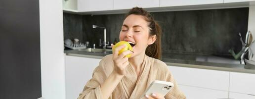 ritratto di felice, bellissimo donna sorridente, mangiare un Mela nel il cucina, seduta a casa nel accappatoio, Tenere smartphone, utilizzando mobile Telefono foto