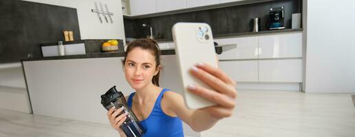 ritratto di giovane fitness ragazza, prende autoscatto su smartphone con acqua bottiglia, bevande acqua durante allenamento, formazione a casa, record gli sport video blog foto