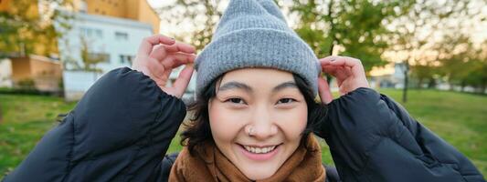 ritratto di carino asiatico ragazza nel cappello e sciarpa, passeggiate in giro cittadina nel freddo primavera tempo atmosferico, sorrisi e sembra felice, si siede nel parco vicino verde erba foto